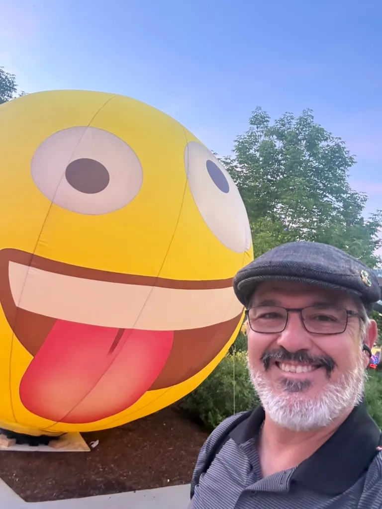 Scott Novis grinning in a flat cap beside a giant inflatable emoji balloon at dusk.