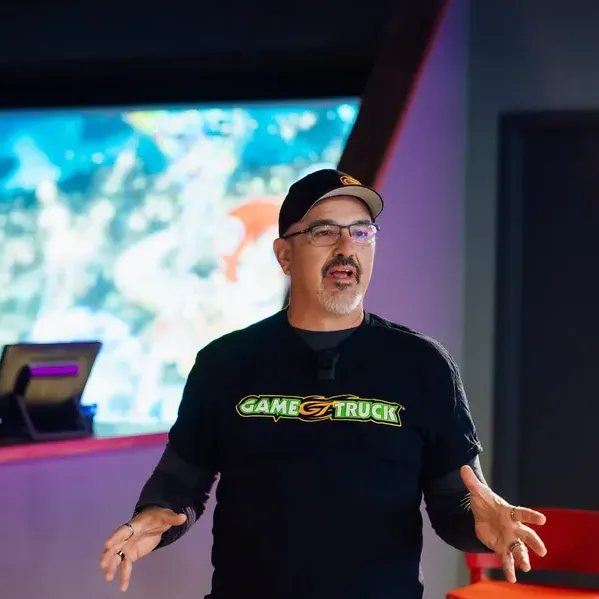Scott Novis speaking on stage in a GameTruck shirt, gesturing mid-talk in front of a large game screen.