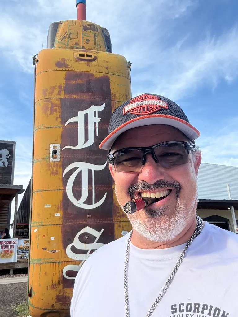 Scott Novis in sunglasses and a Harley-Davidson cap, smiling with a cigar in front of a rusted 'FTW' tank.